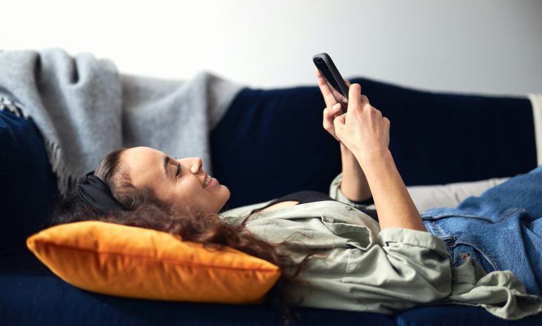 Young Smiling Woman Relaxing At Home Lying On Sofa Checking Social Media On Mobile Phone